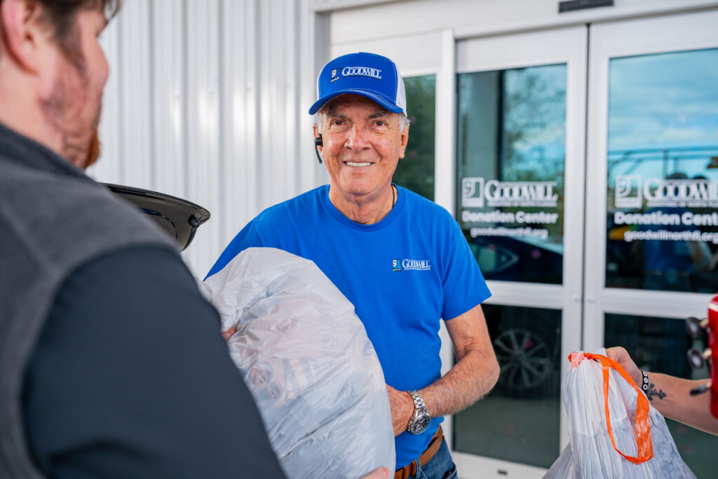 A Goodwill employee in a blue shirt accepts a donation in a white bag in front of a Goodwill donation center.